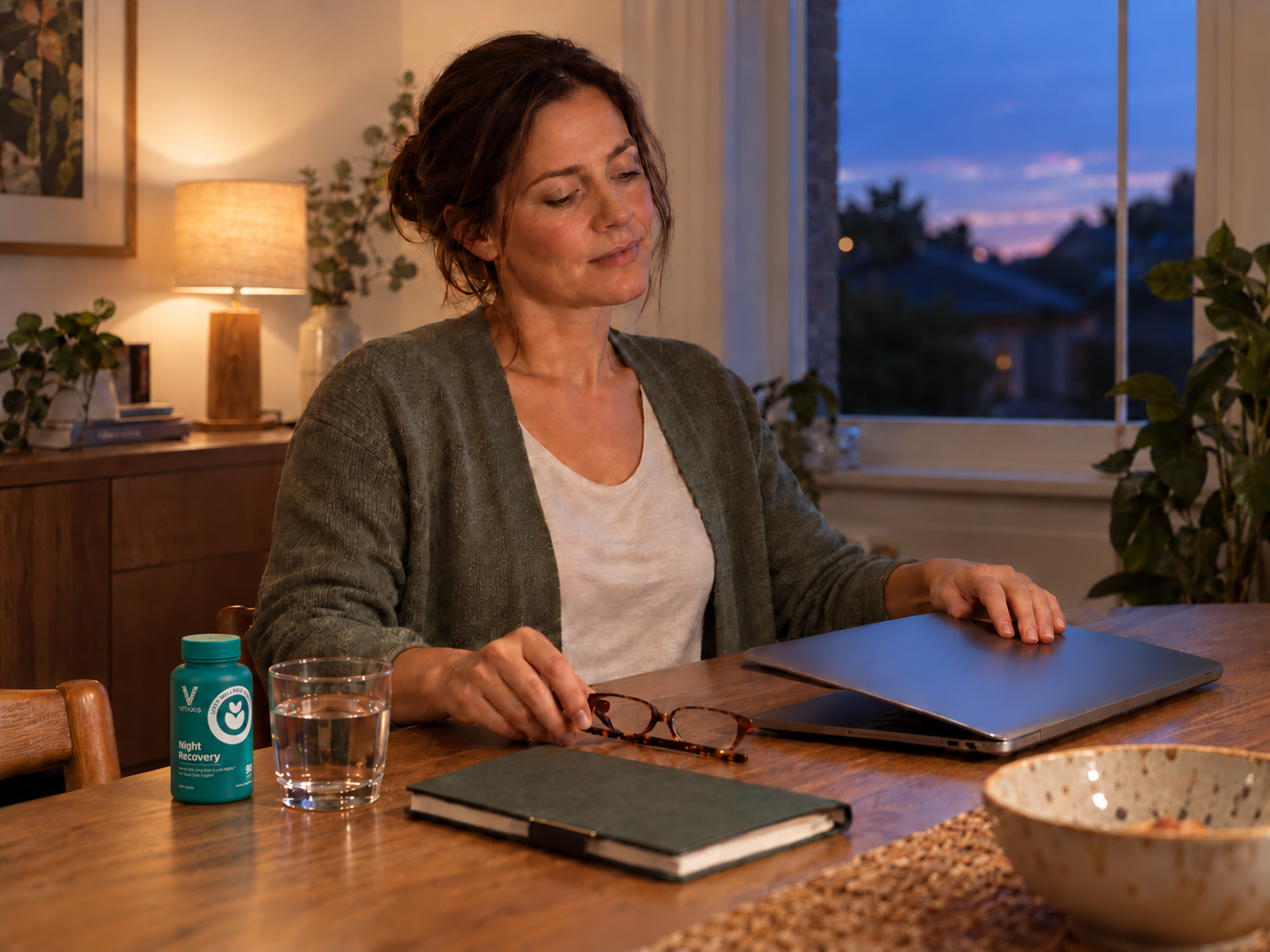A woman finishing work at home with VITAXIS Night Recovery beside her water glass