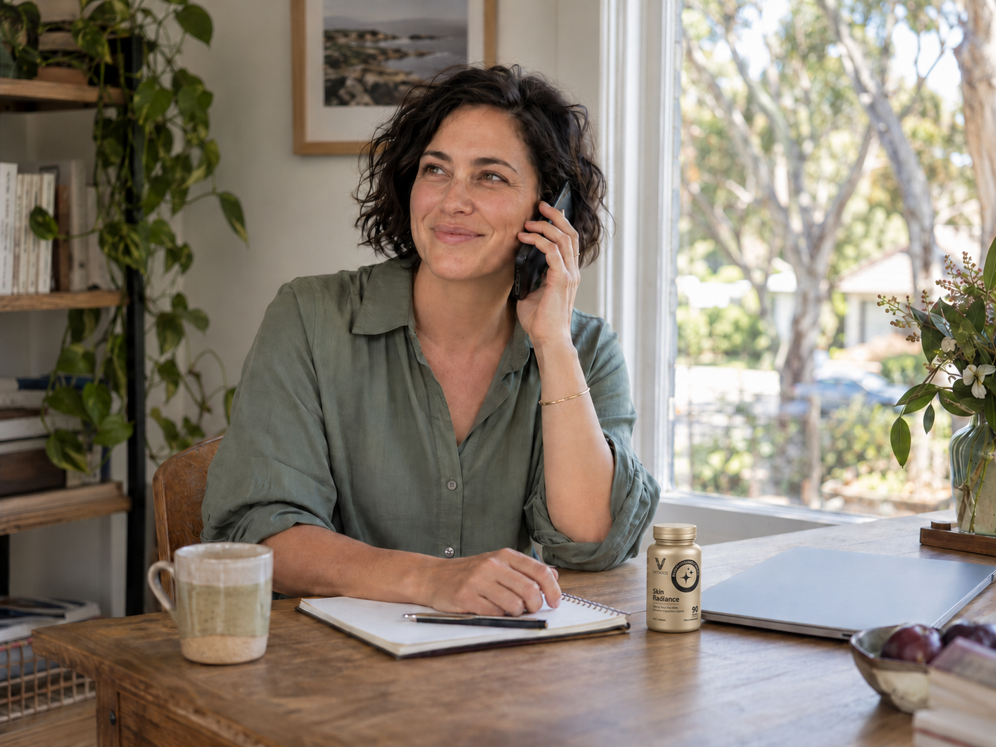 A woman on a phone call with a small VITAXIS Skin Radiance bottle beside her laptop